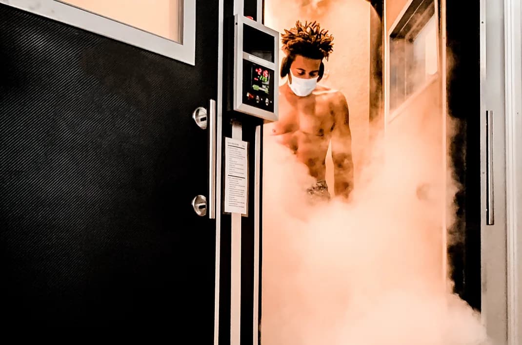 A client in a face mask stepping out of the cryochamber as fog pours across the doorway